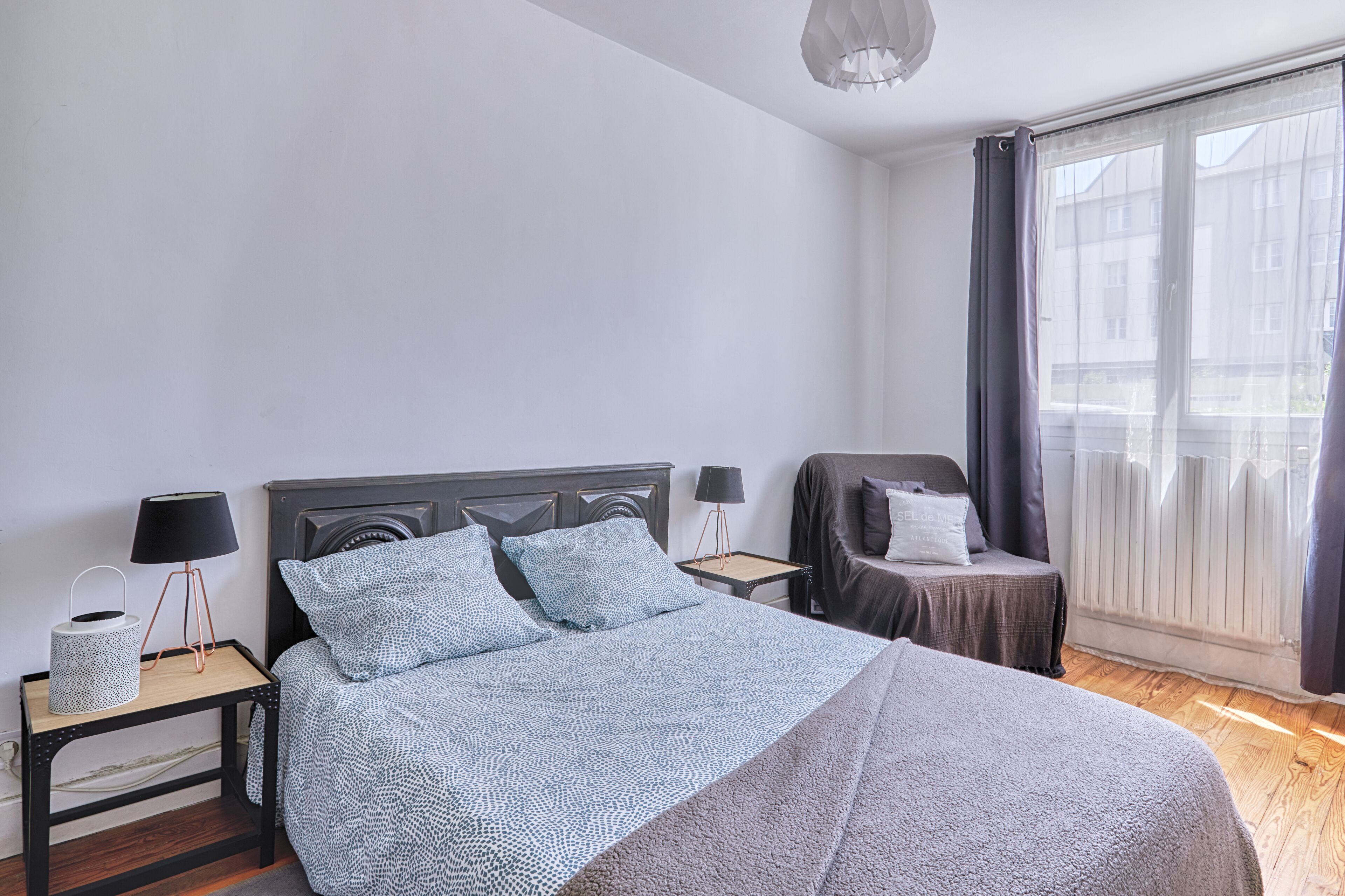 Un dormitorio ordenado con una cama grande, dos mesitas de noche con lámparas, una silla y vistas al exterior a través de una ventana con cortinas.