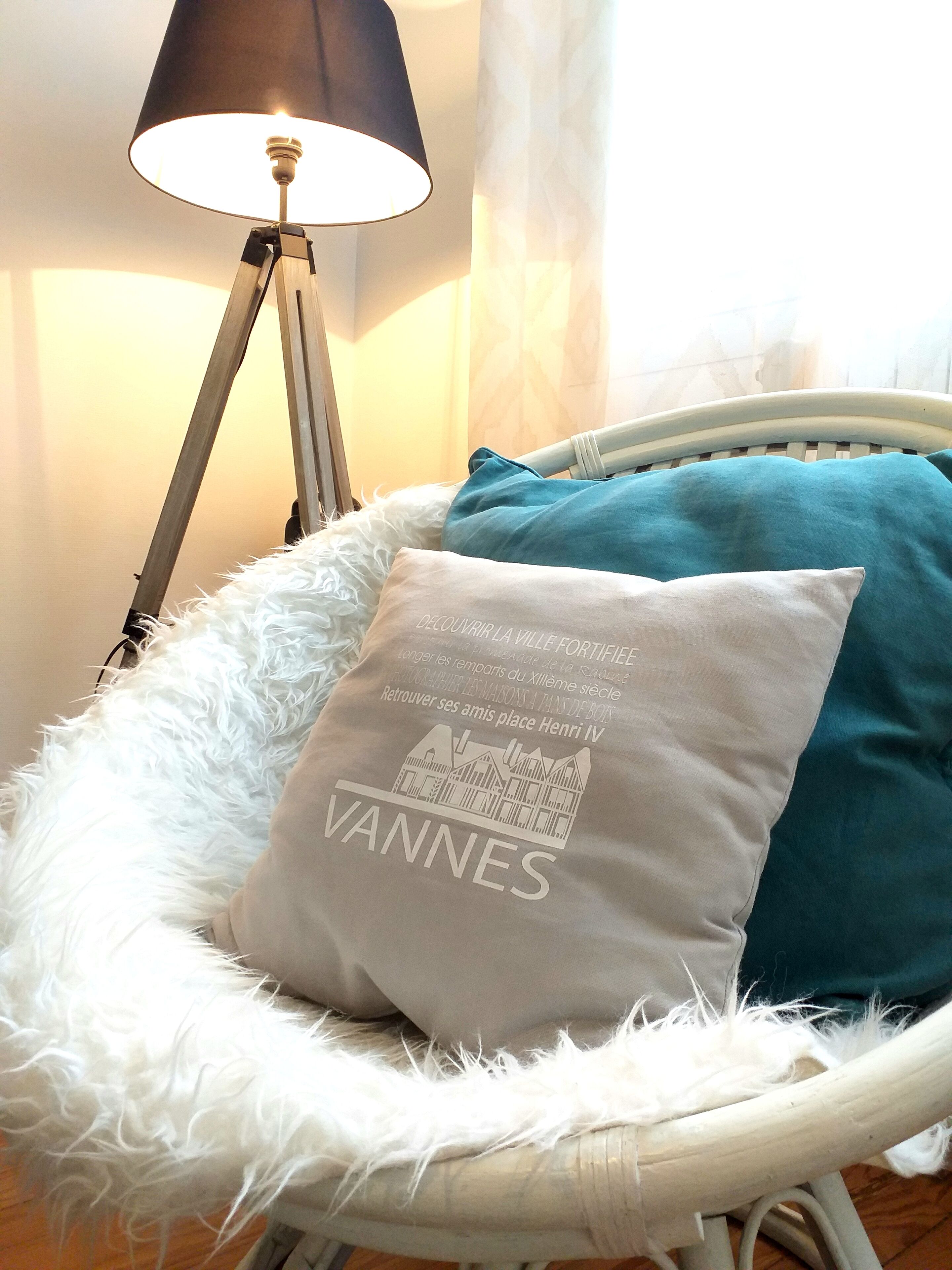 A white wicker chair with a fluffy white cushion and a teal pillow. A lamp with a black shade is positioned behind the chair.