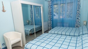 Ein Schlafzimmer mit einem großen Spiegel, einem weißen Stuhl, zwei Betten mit blauer Bettwäsche und einem Fenster mit blauen Vorhängen.