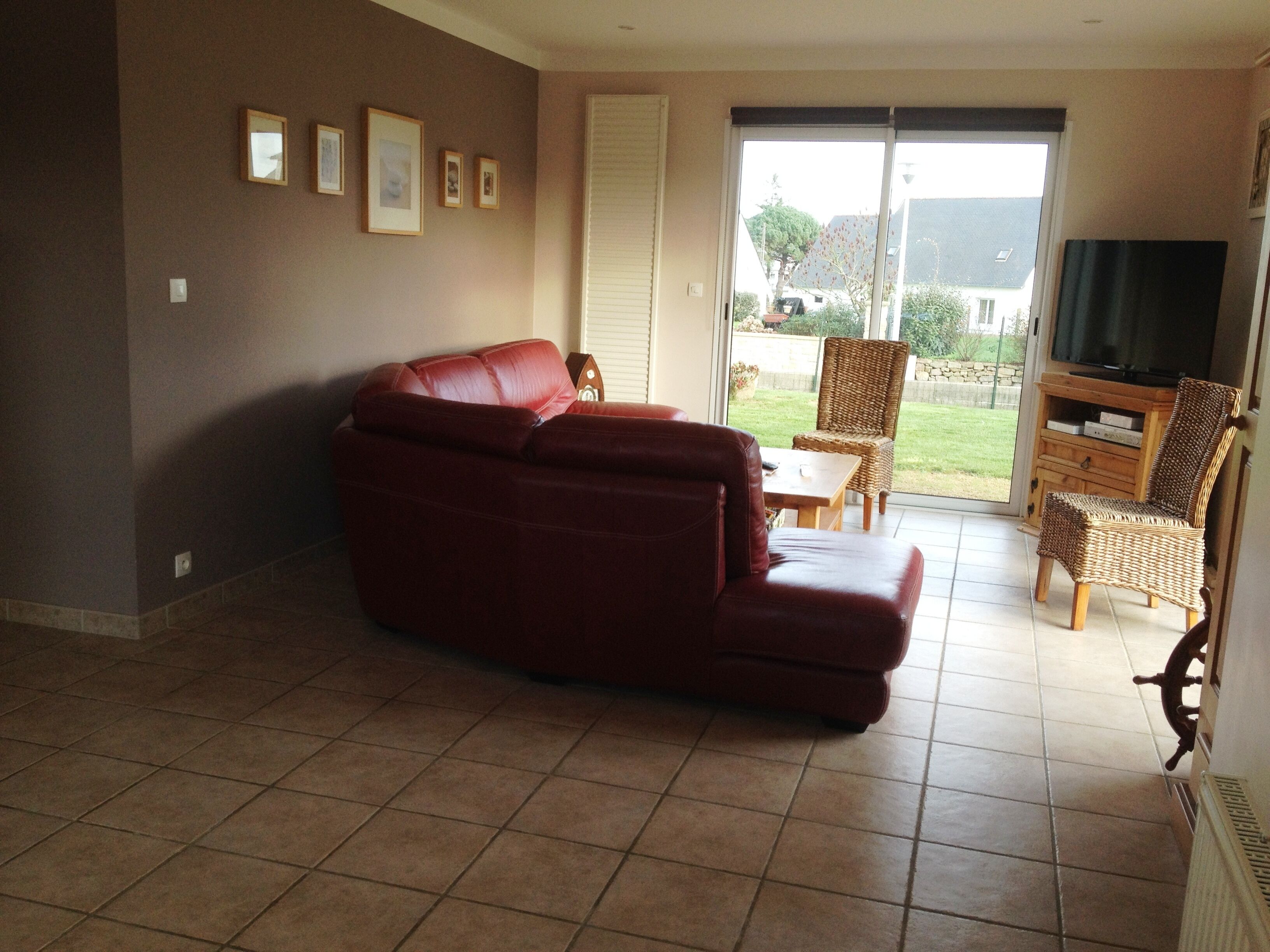 Un salon avec un canapé en cuir rouge, un fauteuil en osier, un meuble TV en bois et une porte-fenêtre coulissante donnant sur un balcon.