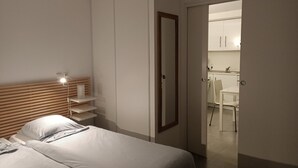 A bedroom with a bed, bedside table, lamp, and wardrobe. A door leads to a small kitchen area with a table and chairs.