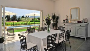 Un coin repas avec une table et des chaises, un vase de fleurs, un meuble contenant des objets décoratifs et une vue sur une piscine et un jardin.