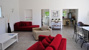 Un salon moderne avec des canapés rouges, un canapé beige et une table basse blanche. On y trouve également un coin repas avec une table et des chaises, et une cuisine à l’arrière-plan.