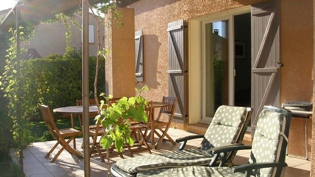 Un patio exterior con mesa y sillas, un espacio de estar cubierto con sillones, y un ventanal corredero que da a un acceso techado.
