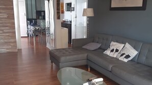 A modern living room with a grey sofa, a glass coffee table, and a dining area in the background.