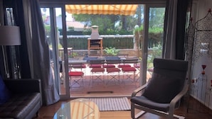 A living room with a glass coffee table, a sofa, and a chair. There is a sliding glass door leading to an outdoor patio with a table and chairs.