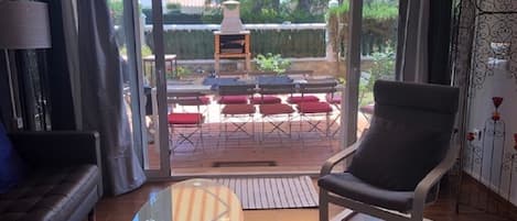 Una sala de estar con una mesa de centro de vidrio, un sofá y una silla. Hay una puerta corrediza de vidrio que da a un patio exterior con una mesa y sillas.