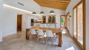 Un comedor amplio con una mesa y sillas de madera, una cocina al fondo y ventanas grandes que dejan entrar luz natural.