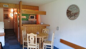 Een kleine keuken met een magnetron, houten kastjes en een eethoek met een tafel en stoelen.