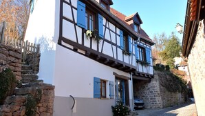 A traditional half-timbered house with a stone staircase leading to the entrance, a car parked outside, and a clear sky.