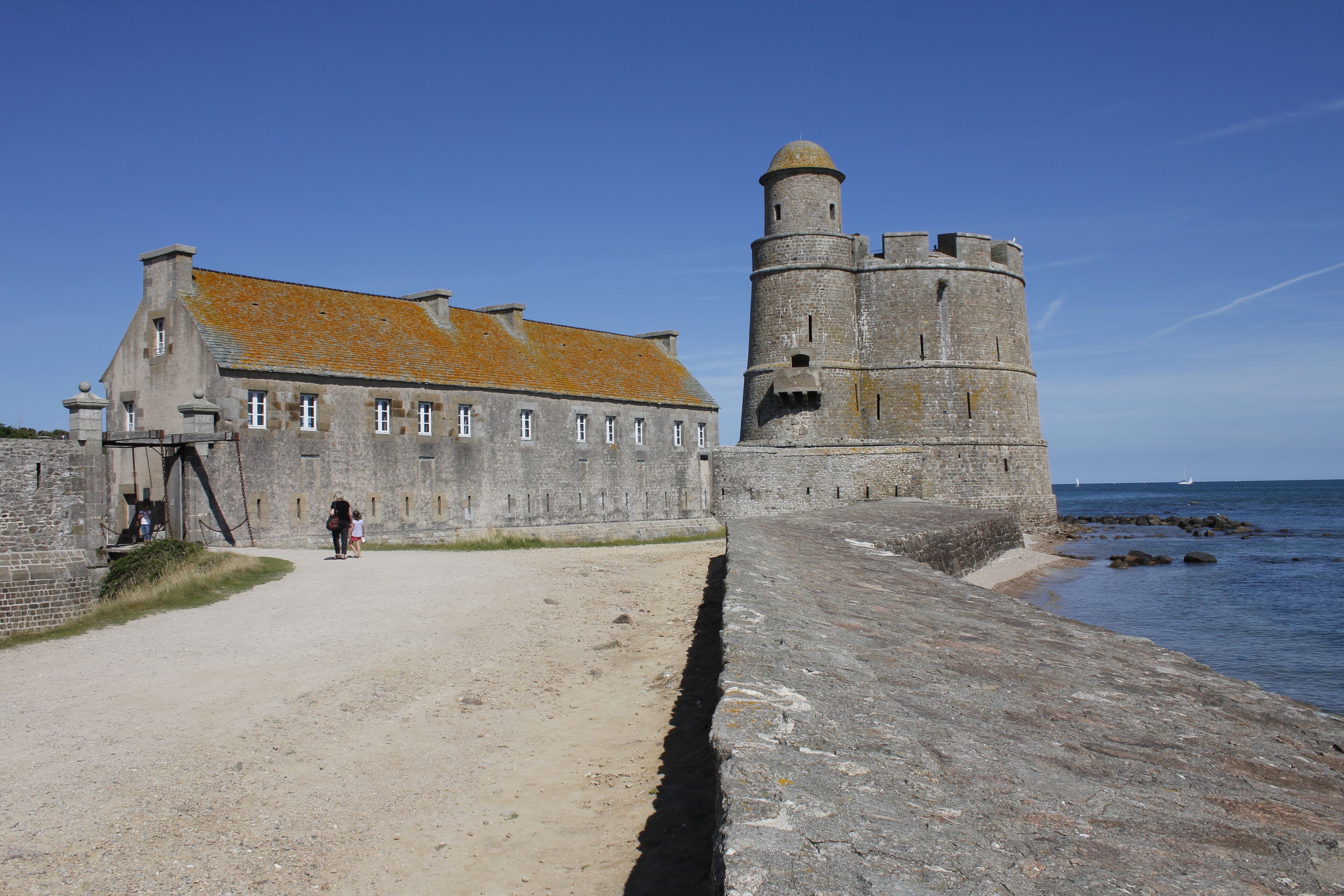 Ein historisches Steinhaus mit einem Turm, einem Feldweg und einem Gewässer.