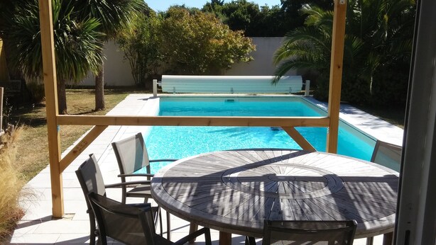 Een terras buiten met een zwembad, een pergola en een ronde tafel met stoelen.