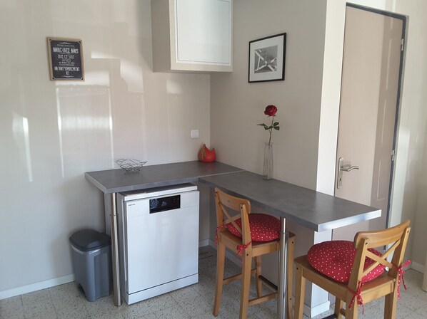 A compact kitchen with a dishwasher, two wooden chairs with red cushions, a small trash bin, a framed quote on the wall, and a vase with a single red rose.