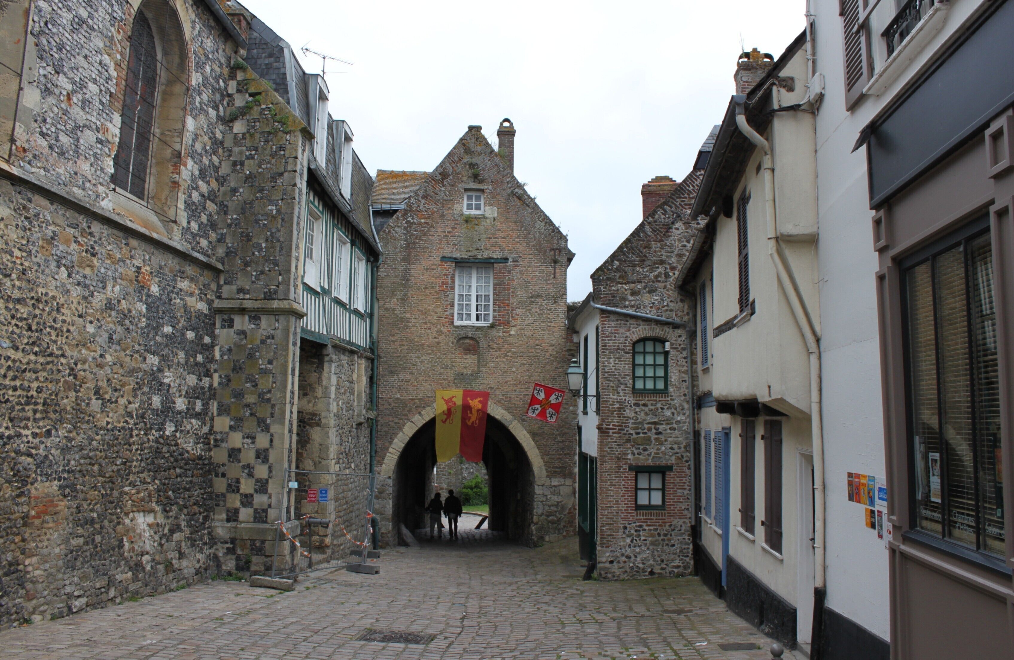 A narrow cobblestone street with historic stone buildings, a stone archway, and flags.
