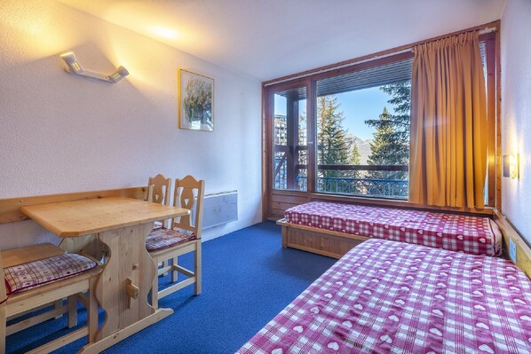A room with a wooden table and bench, two beds with checkered bedding, a window with curtains, and a painting on the wall.