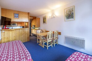 A room with a wooden kitchenette, a dining table with chairs, a bed with a checkered bedspread, and a painting on the wall.