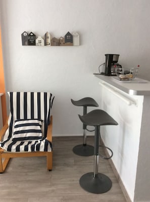 Una pequeña cocina con una silla de madera, dos taburetes de bar y una máquina de café sobre una encimera blanca.