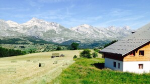 Une maison de campagne avec un toit en pente, entourée d’un champ verdoyant et bordée de montagnes en arrière-plan.