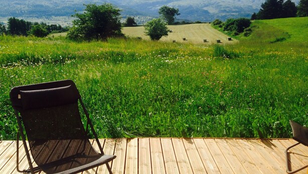 Une terrasse en bois avec une chaise en métal, donnant sur un champ verdoyant avec des montagnes en arrière-plan.