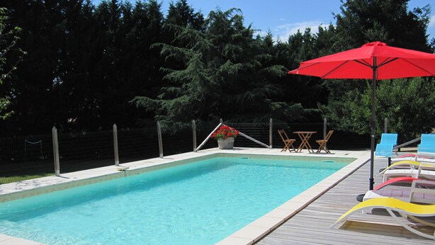 Una piscina rectangular con una solana empavedada, rodeada por una cerca y árboles. Hay sillas de descanso y una sombrilla roja cerca de la piscina.