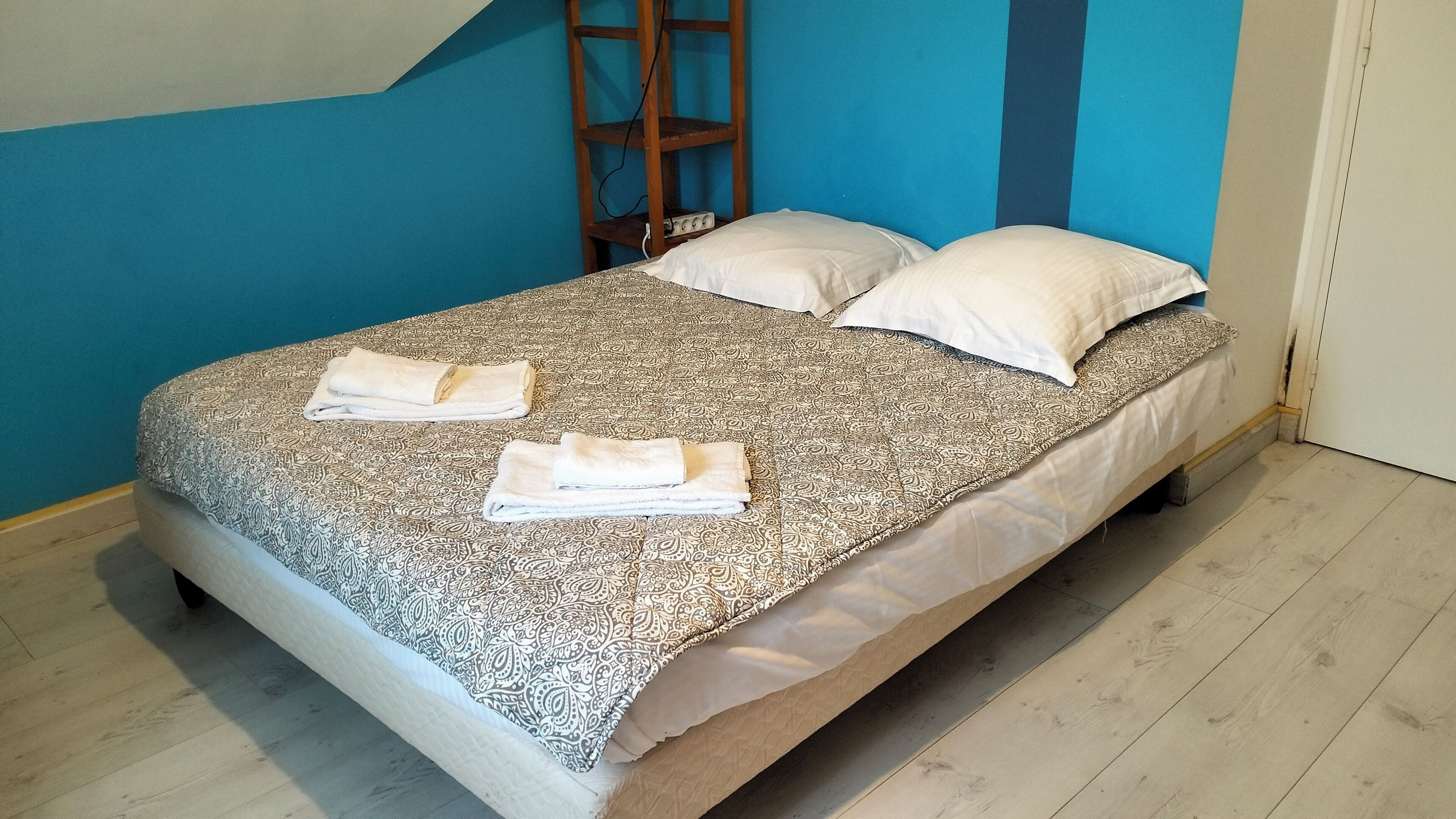 A single bed with a patterned bedspread and two pillows, a wooden ladder against the wall, and a light-colored wooden floor.