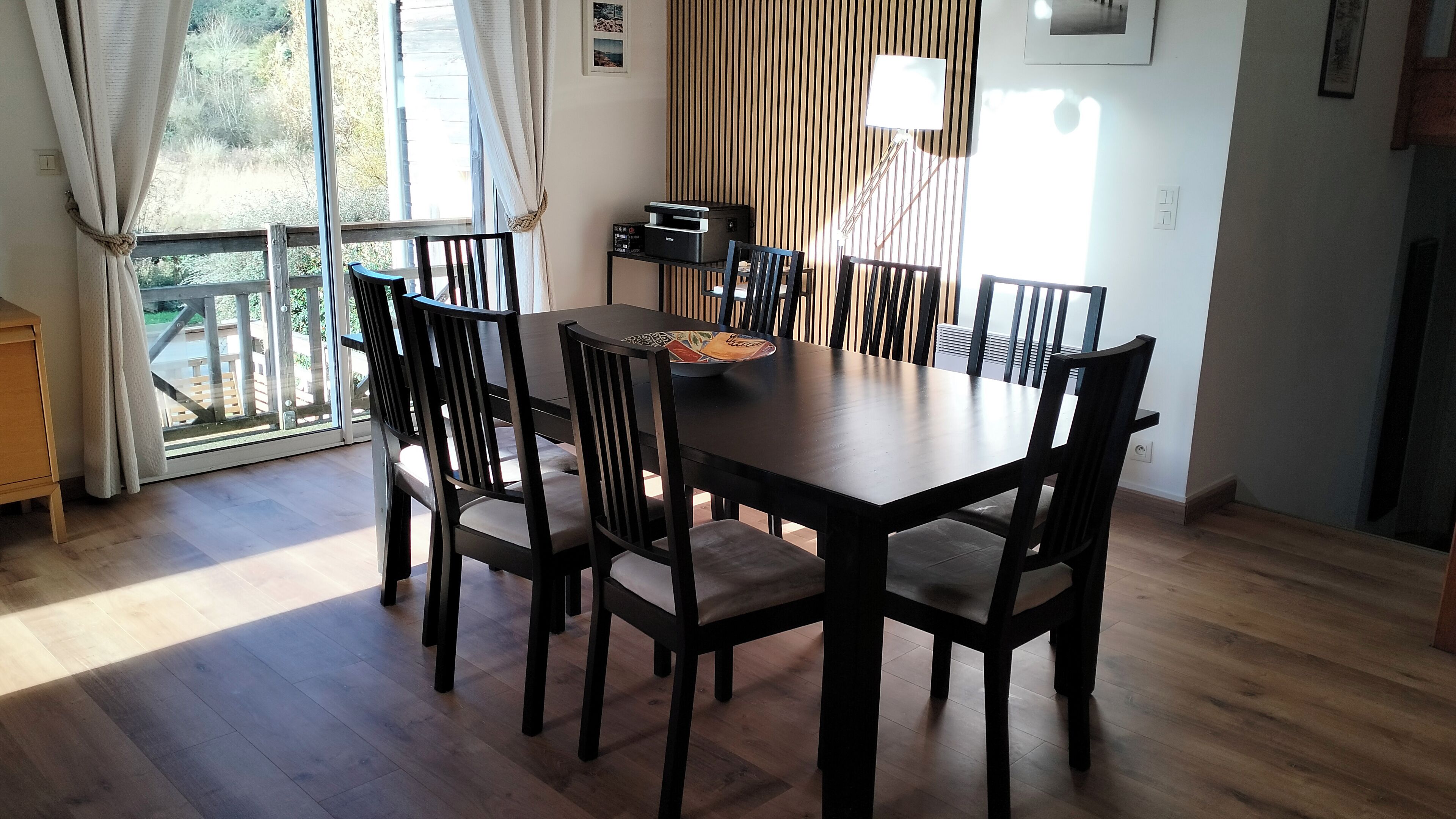 A dining area with a wooden table and chairs, a printer on a small table, and a view of greenery through the window.