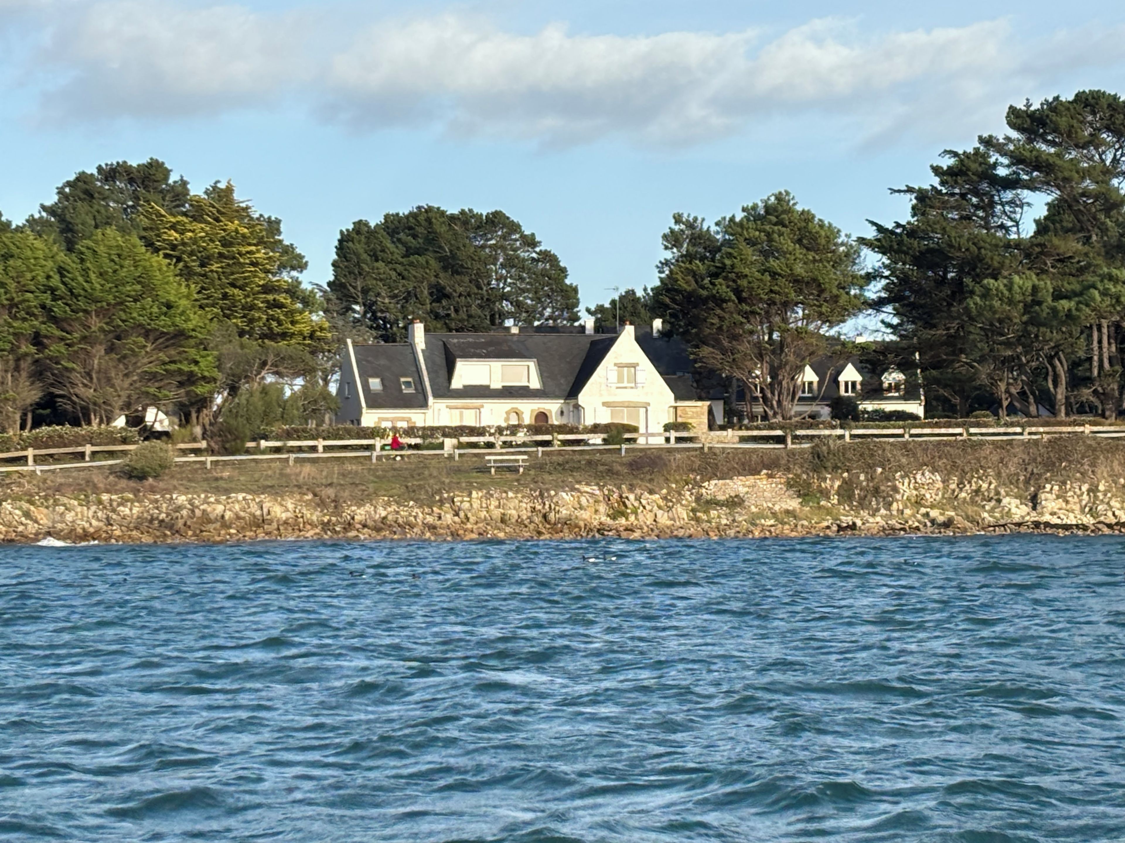 A coastal house with a stone wall and a wooden fence, overlooking a body of water.
