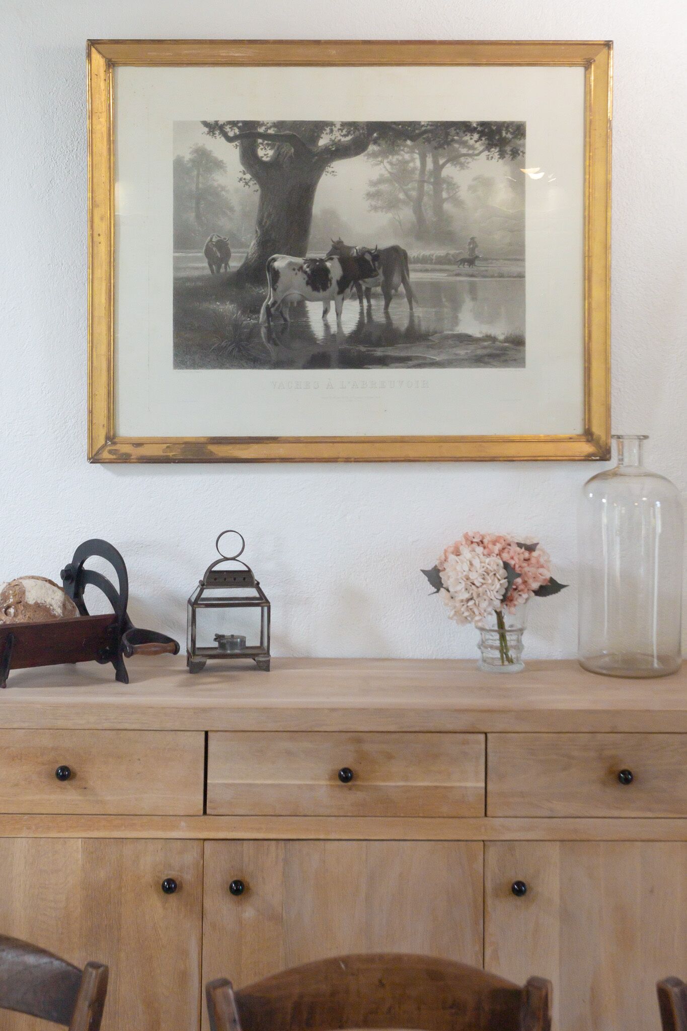Une photographie encadrée représentant des vaches près d’un arbre, une lanterne, un vase de fleurs et une bouteille en verre posés sur une commode en bois.