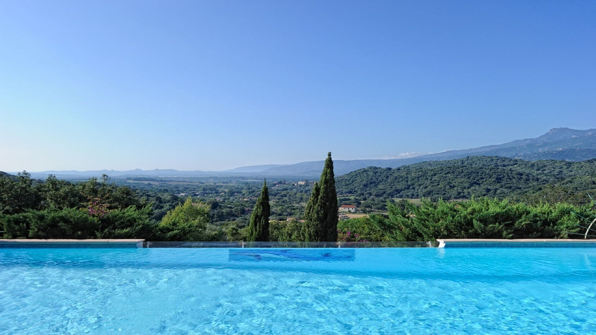 Une piscine à l’eau d’un bleu cristallin, entourée d’une végétation luxuriante et de grands cyprès, offrant une vue imprenable sur un paysage montagneux.