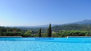 A swimming pool with clear blue water, surrounded by lush greenery and tall cypress trees, overlooking a mountainous landscape.