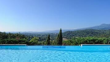 Une piscine à l’eau d’un bleu cristallin, entourée d’une végétation luxuriante et de grands cyprès, offrant une vue imprenable sur un paysage montagneux.