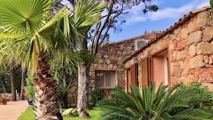 A stone building with a tiled roof, a window with shutters, and a well-maintained garden with palm trees and other greenery.