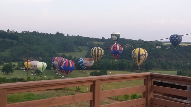 Vue sur des montgolfières survolant un paysage verdoyant, prise depuis un balcon en bois.