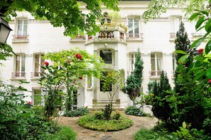 A white building with a central entrance, surrounded by a garden with various plants and a pathway leading to the entrance.