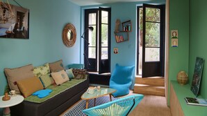 A cozy living room with a green sofa, a blue armchair, and a small coffee table. There is a door leading to a bright room with a view of trees. - atypical design studio, 2 terraces (Paris)
