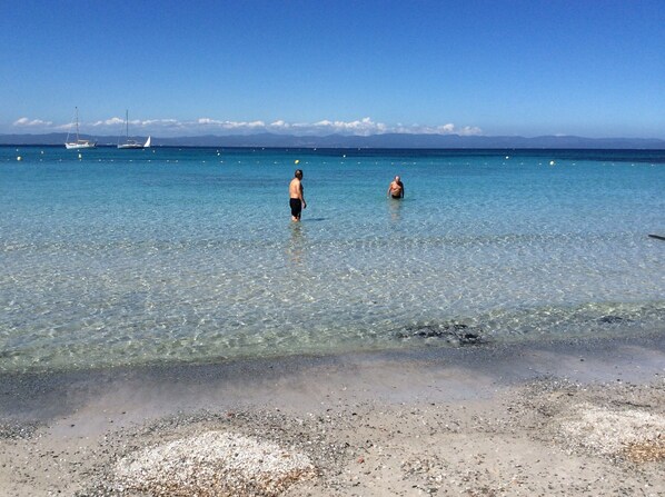 A beach with clear water, pebbles, and distant boats.
