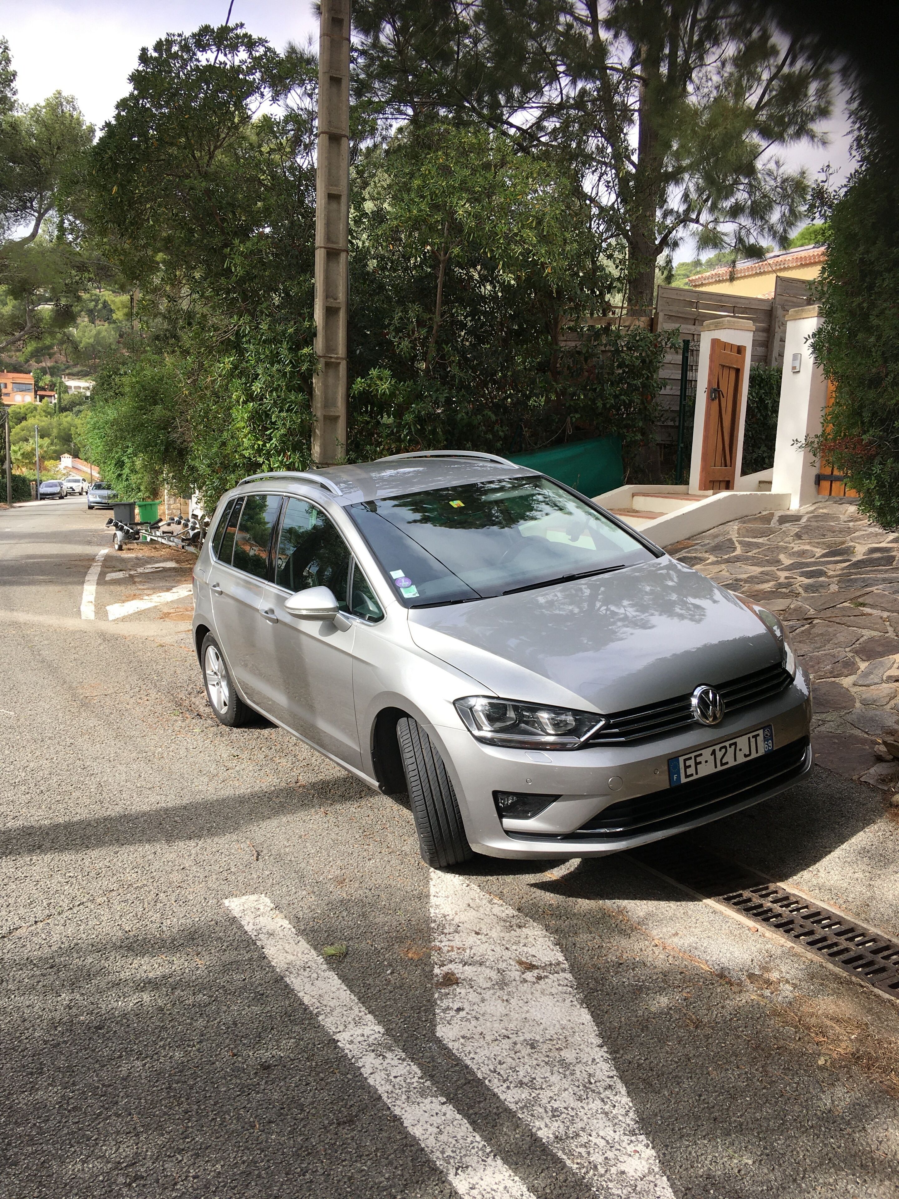 En silverfärgad Volkswagen-bil parkerad på en gata, med en stenmur och en trädörr i bakgrunden.