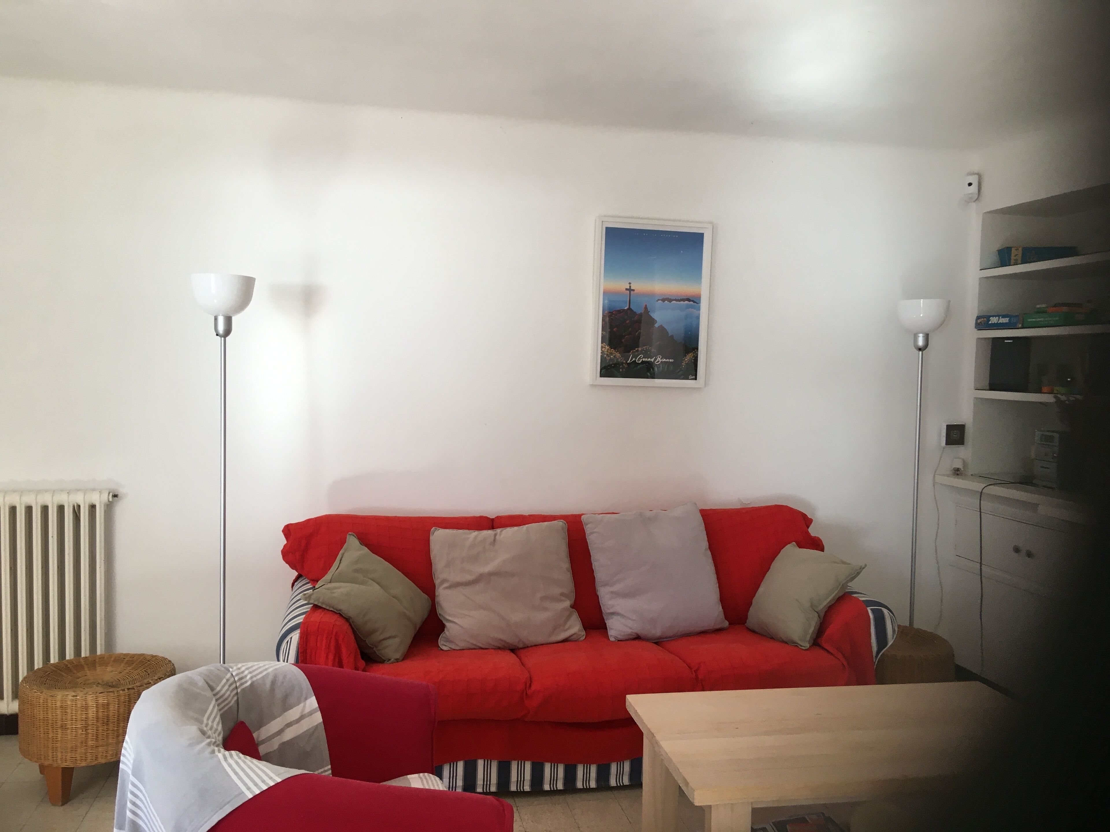 A red sofa with striped cushions, a wicker chair, a wooden table, and a white radiator.