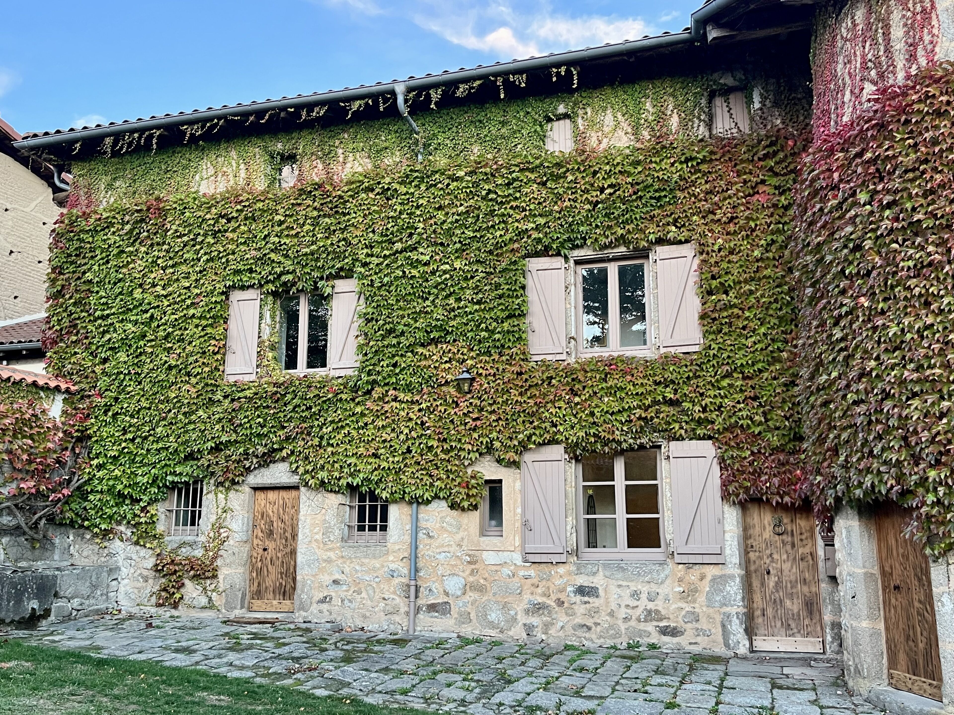 Un bâtiment en pierre aux murs recouverts de lierre, doté de volets en bois et d’une cour pavée.
