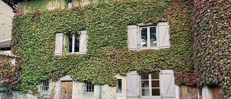 Un bâtiment en pierre aux murs recouverts de lierre, doté de volets en bois et d’une cour pavée.