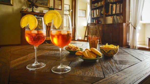 Two glasses of a red beverage with lemon slices, a bowl of breadsticks, and a bowl of chips on a wooden table in a room with bookshelves and a painting. - Charming apartment in the historic center (Roma)
