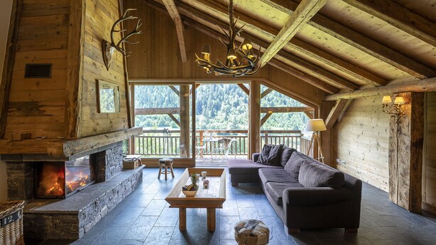 A log cabin interior with a stone fireplace, a wooden ceiling, and a large window overlooking a forest. - Pags Haut, Pags Haut (Châtel)
