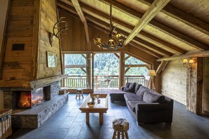 Interior d'una cabana de fusta amb una llar de pedra, un sostre de fusta i una finestra gran amb vista a un bosc.