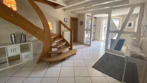 A spacious living area with a wooden staircase, white tiled flooring, and a view of the outdoors through glass doors.