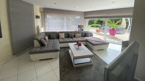A modern living room with a sectional sofa, a coffee table, and a TV. There is a large window providing a view of the outdoor patio area.