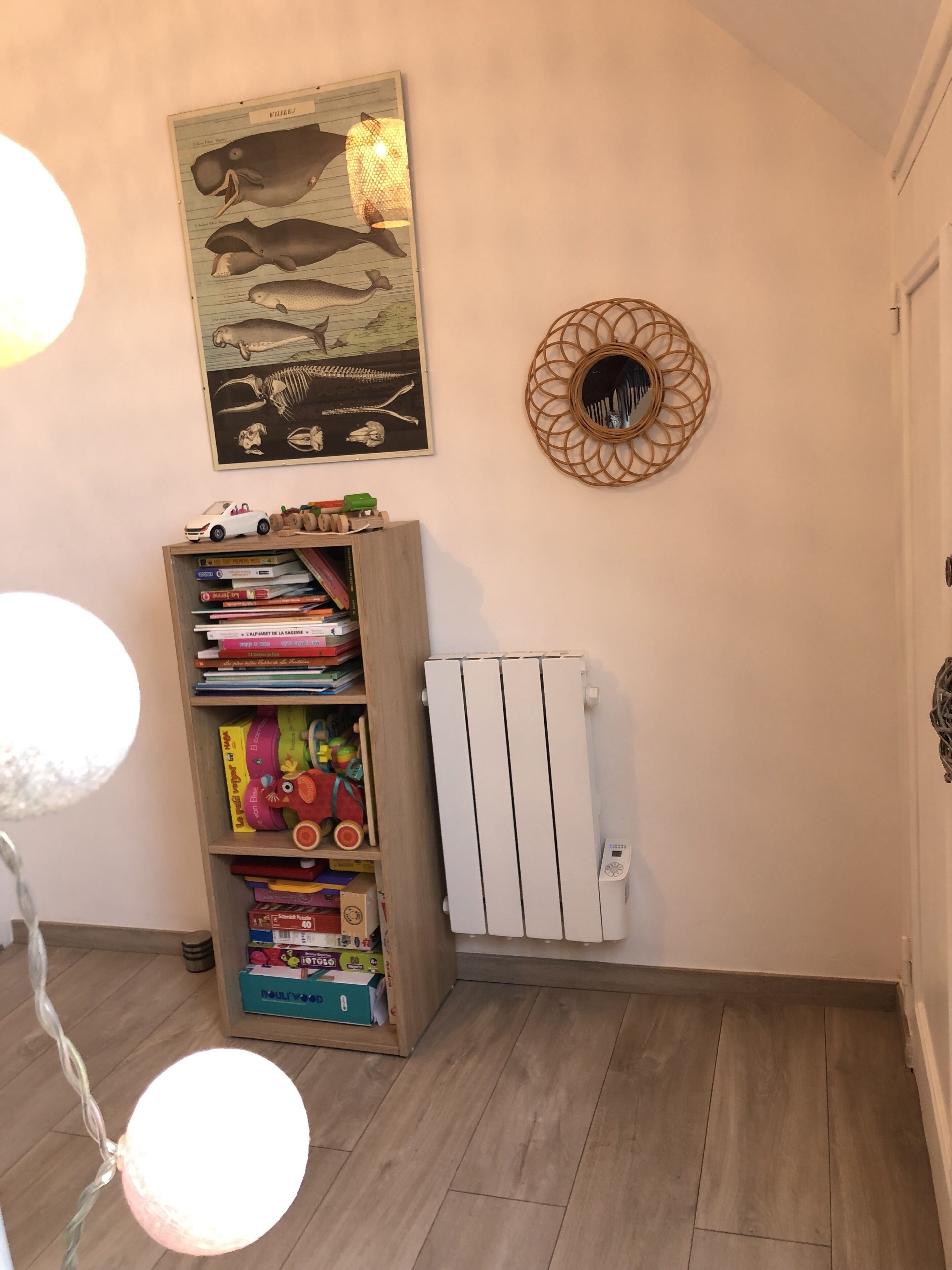 Et værelse med en træbogreol fyldt med bøger og legetøj, en radiator og en dekorativ genstand på væggen.