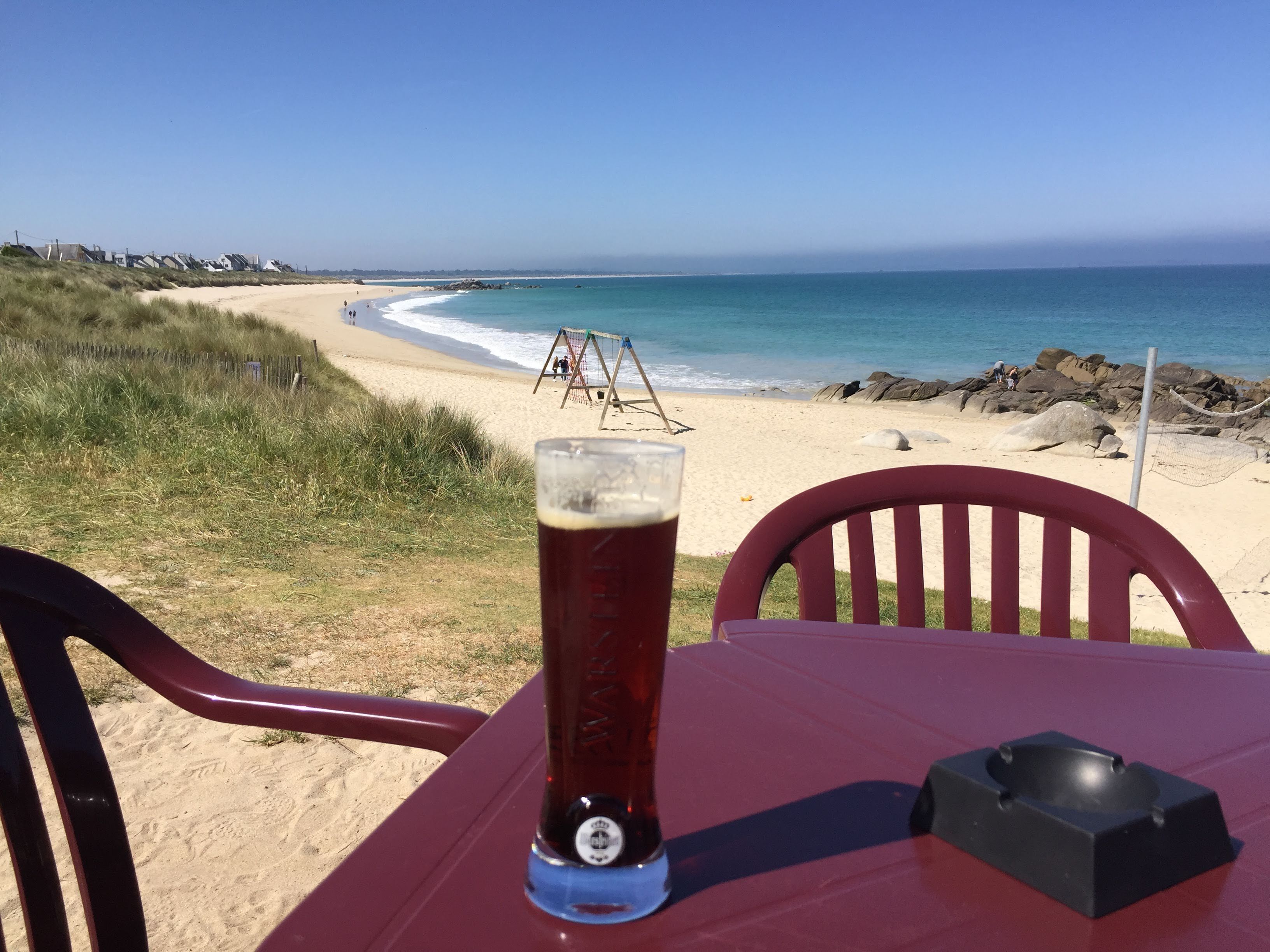 A table with a glass of beer and a black ashtray, overlooking a beach with a volleyball net and rocky shoreline.