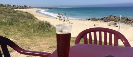 A table with a glass of beer and a black ashtray, overlooking a beach with a volleyball net and rocky shoreline.