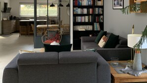 Un salon moderne avec un canapé d’angle gris, une table basse en bois, une bibliothèque et un coin repas séparé du reste de la pièce par une cloison en verre, d’où l’on aperçoit la cuisine.