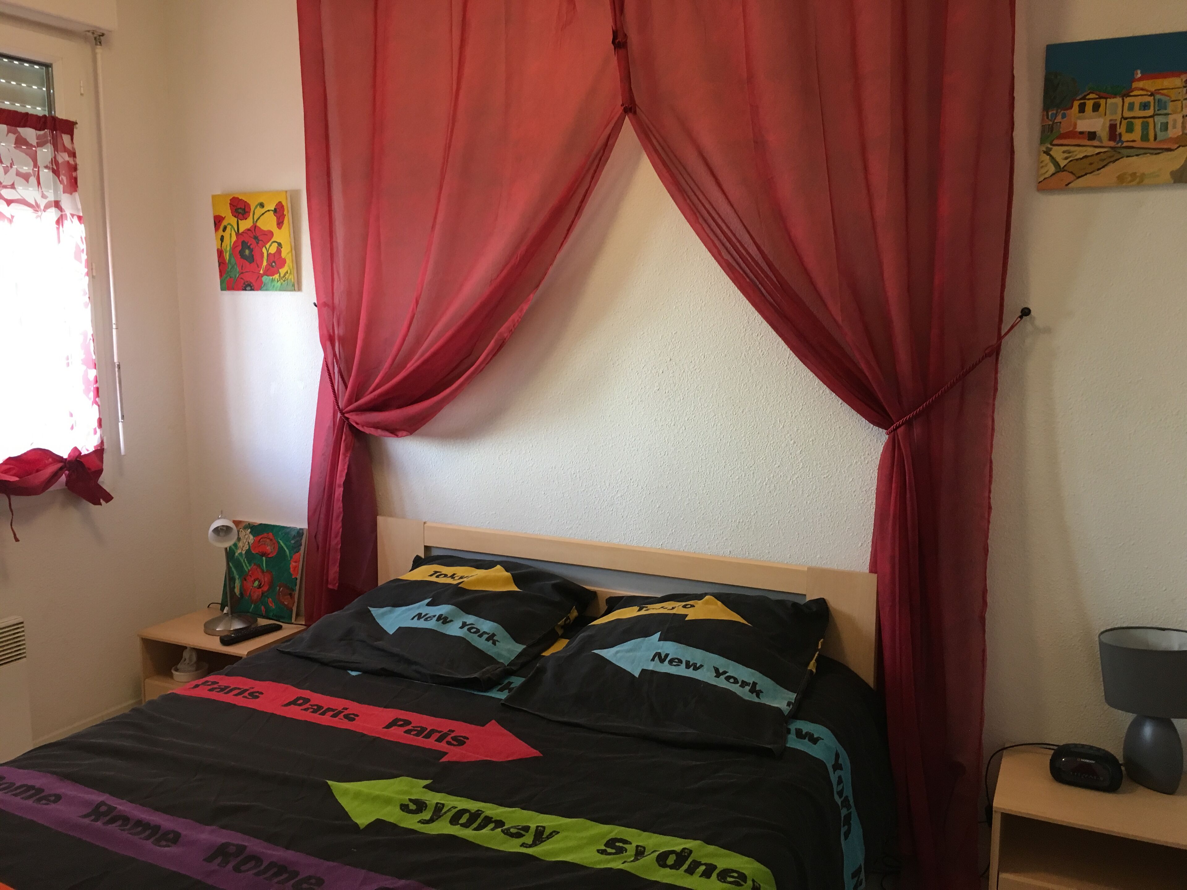 Une chambre à coucher avec un lit orné de flèches directionnelles pointant vers différentes villes, des rideaux rouges, une table de chevet avec une lampe et un tableau mural.